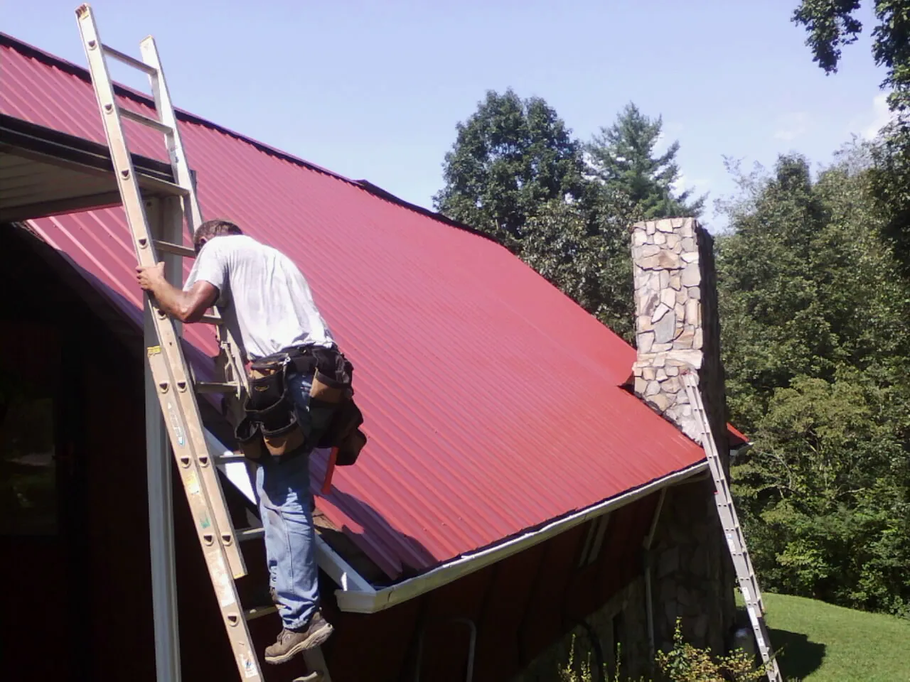 Roofer on a ladder beside a red metal roof and stone chimney — Soffit & Fascia Repair by Roofing Brothers in Maple Valley